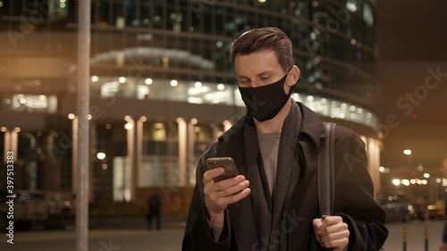 Man outdoors wearing a black protective mask, looking at the phone. Man in black clothes standing on background of city buildings and road with cars