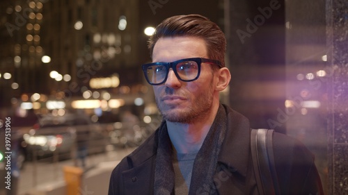 Man in eyeglasses standing on background of business skyscrapers, windows with lights. Portrait gimbal shot of businessman in coat standing near business centre downtown at night