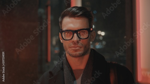 Man in eyeglasses looking at the camera with red lights on face, night time. Portrait shot of serious businessman outside on background of glass window