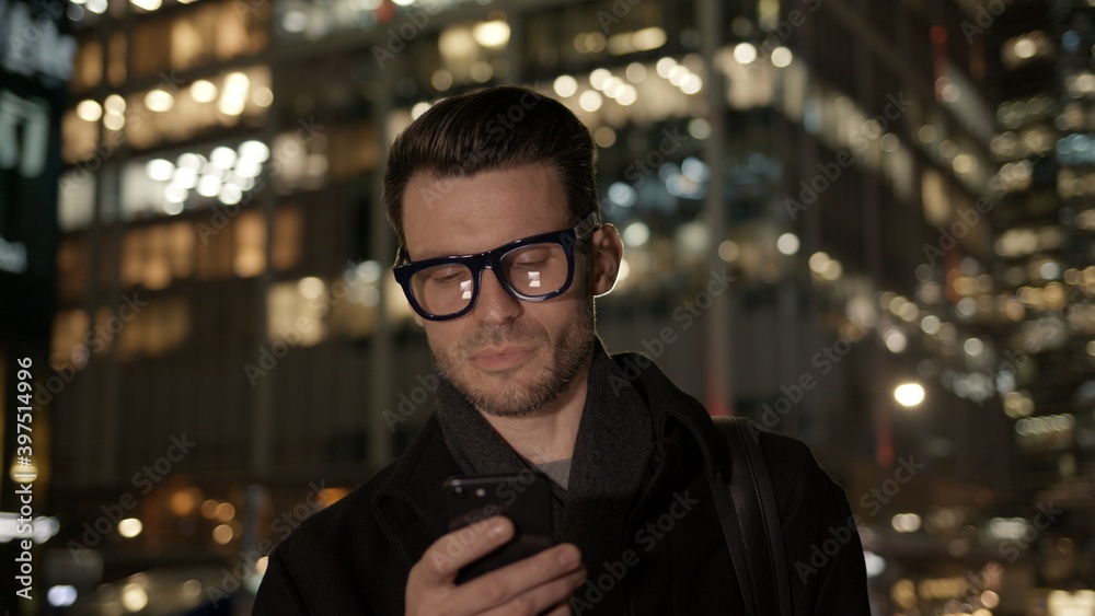 Man in eyeglasses with phone in hand on background of building with lights on. Gimbal night shot of caucasian man texting in phone near business building