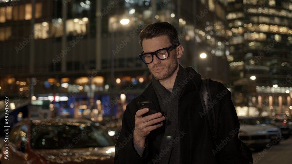 Man in eyeglasses with phone in hand on background of building with lights on. Gimbal night shot of caucasian man texting in phone near business building