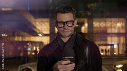 Man in eyeglasses with phone in hand on background of building with lights on. Gimbal night shot of caucasian man texting in phone near business building