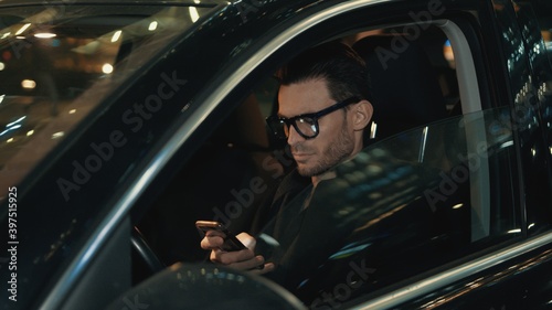 Businessman sitting in car with phone in hand, man in eyeglasses on a driver seat typing looking at the phone. Handheld shot of business taxi driver at night time