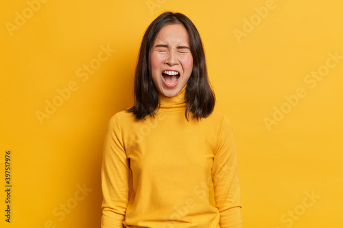 Emotional dark haired Asian woman screams loudly expresses rage keeps mouth widely opened wears casual yellow turtleneck in one tone with studio background. Human emotions and feelings concept