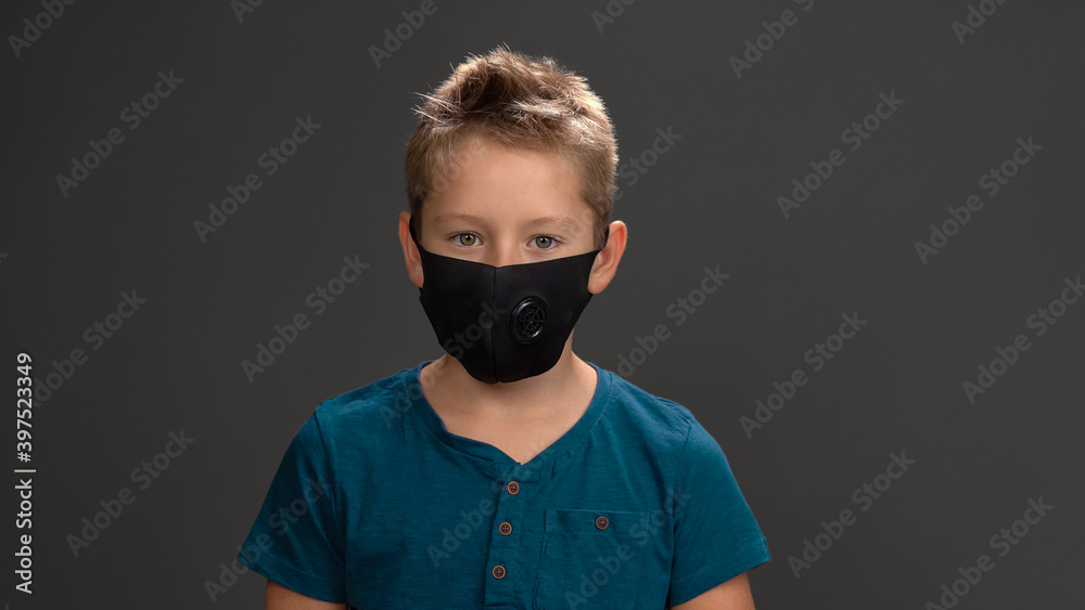 Cute schoolboy in face reusable mask, dark blue t-shirt smiling at camera while standing in studio isolated on black background. Fashion concept. Cute schoolboy in face reusable mask, dark blue t-shirt smiling at camera while standing in studio isolated on black background. Fashion concept.