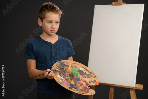 Artist little boy in blue t-shirt looking sad holding a palette with paints dried upstanding next to an canvas on the easel. School of arts concept.  photo