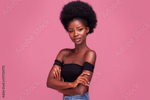 Confident African American girl with afro hair standing light smiling with arms folded in black bare shoulder top and blue denim jeans isolated on pink background.  photo