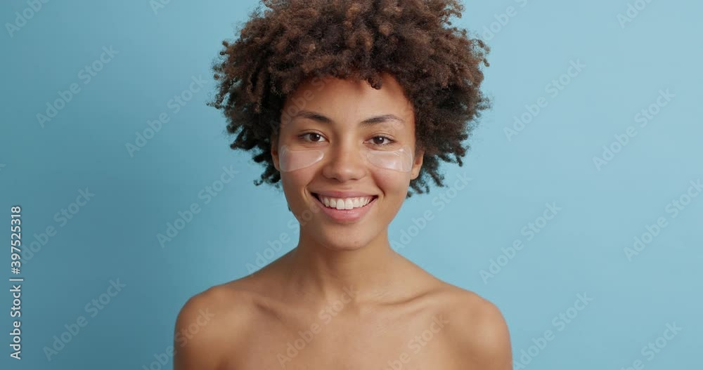 Positive dark skinned curly young woman with naked body smiles positively has good mood curly hair enjoys skin care procedures isolated over blue background. Cosmetology and wellness concept Positive dark skinned curly young woman with naked body smiles positively has good mood curly hair enjoys skin care procedures isolated over blue background. Cosmetology and wellness concept