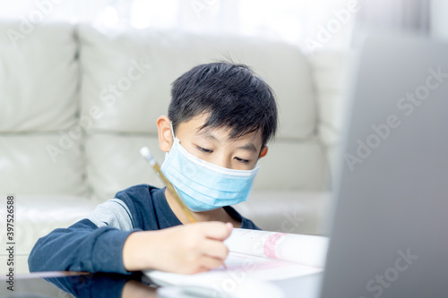 asian boy doing homework with computerlaptop at home. Child using gadgets to study.Education and distance learning for kids. Homeschooling during quarantine. Stay at home entertainment.child with mask photo
