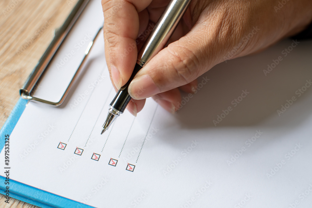 Close- up, Men's fingers holding pen writing on checklist paper and the format for filling in information in business concept. Businessman writing on checklist on wooden desk. Blurred wood background Close- up, Men's fingers holding pen writing on checklist paper and the format for filling in information in business concept. Businessman writing on checklist on wooden desk. Blurred wood background