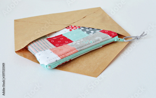 Open kraft envelope and colorful patchwork quilted zipper pouch over white photo