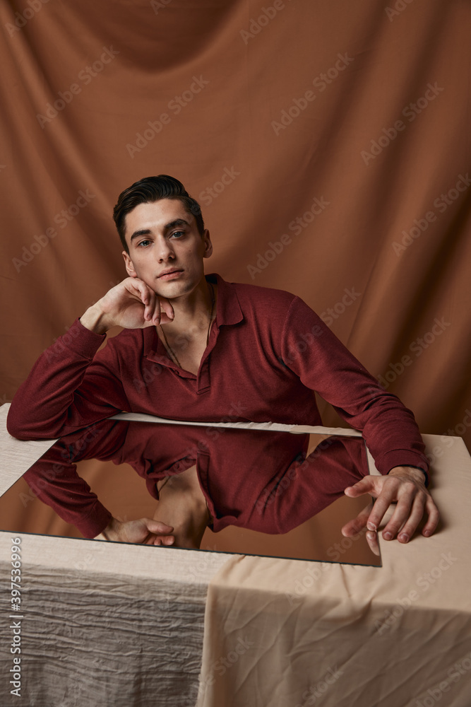 Fototapeta premium Handsome guy in a shirt posing at a table indoors on a fabric background and a square mirror