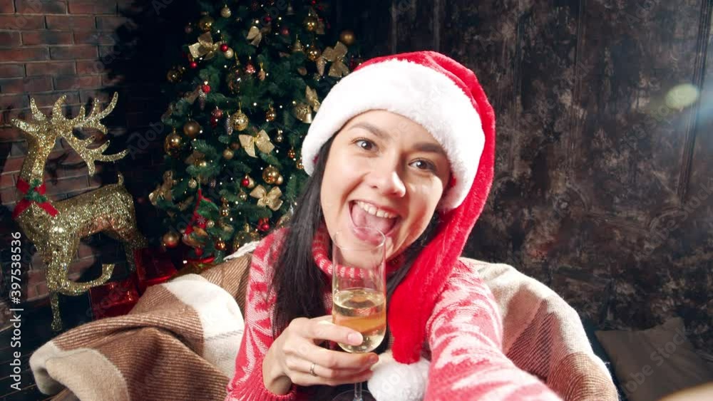 Portrait of a young woman in a Santa hat drinking champagne and taking a selfie or recording a video on the web camera of a smartphone while sitting near a Christmas tree at home Portrait of a young woman in a Santa hat drinking champagne and taking a selfie or recording a video on the web camera of a smartphone while sitting near a Christmas tree at home