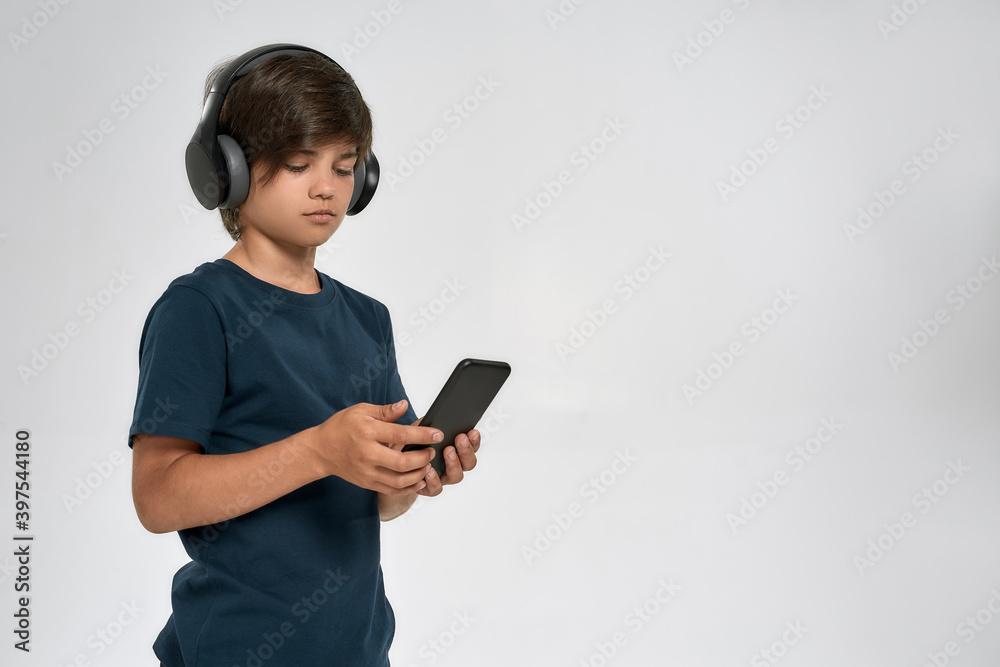 Little sportive boy child in sportswear wearing headphones, using smartphone, standing isolated over white background Little sportive boy child in sportswear wearing headphones, using smartphone, standing isolated over white background
