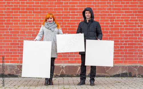 Young man and woman standing against a background of a red brick wall holding three white canvases in front of them to paint