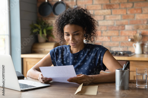 Smiling pleased young african american woman reading paper letter with good news, sitting at table with computer at home. Happy curious pretty millennial mixed race lady getting correspondence.