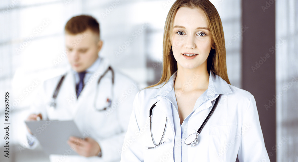 Fototapeta premium Professional beautiful woman-doctor with a stethoscope is standing with crossed arms in a clinic. Young doctors at work in a hospital. Medicine and healthcare concept