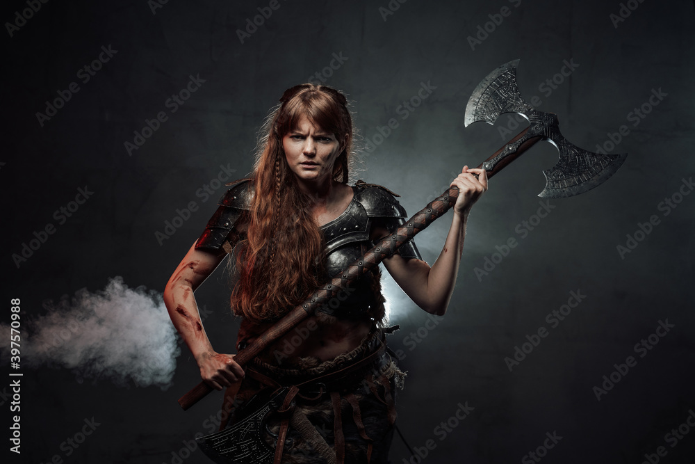 Violent and savage scandinavian female warrior holding huge two handed axe in dark foggy background.