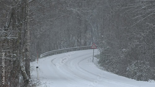 Snow falling on curved mountain countryside road in Slovenia. Cold snowstorm in winter season. Forest surrounding empty road. Static shot, real time, wide angle