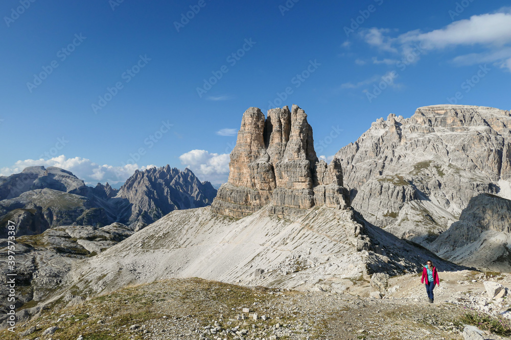 Obraz premium Woman in pink jacket hiking on a narrow path in Italian Dolomites. There are sharp and steep mountains around, with Tobling Knoten behind her. Sunny day. Raw landscape. Freedom and exploration