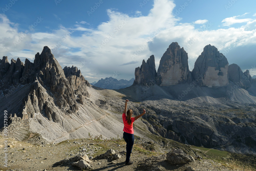 A woman in hiking outfit standing with her arms wide open and enjoying the view on the famous Tre Cime di Lavaredo (Drei Zinnen), mountains in Italian Dolomites. Desolated and raw landscape. Freedom
