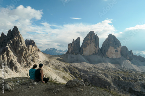 A couple in hiking outfit, sitting on the ground and enjoying the view on the famous Tre Cime di Lavaredo (Drei Zinnen) in Italian Dolomites. Desolated, raw landscape, full of lose stones. Sunny day © Chris