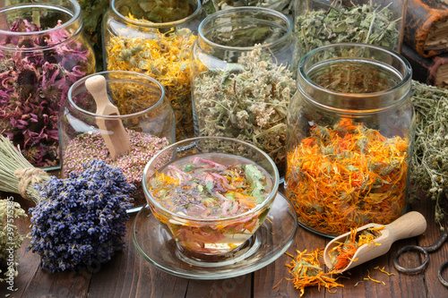 Photography Cup of healthy calendula tea, Jars of dry medicinal herbs - heather, calendula, coneflowers, melissa, linden tree flowers, bunch of dry lavender on wooden table