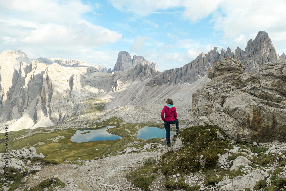 Woman hiking with the view on small, navy blue lakes at the bottom of the valley in Italian Alps. The lakes are surrounded by high and steep peaks The sky is full of soft clouds. Raw landscape. Remedy