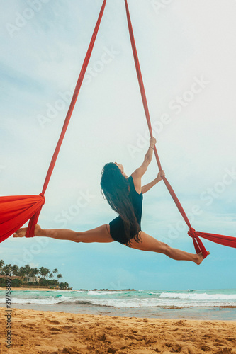 girl aerialist in black swimsuit makes aerial trick on the red airsilk on the palm tree on the sky background, sport art concept, free space 