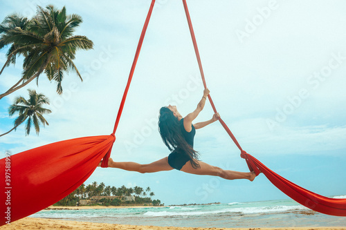 woman aerialist in black swimsuit makes aerial trick  on the red airsilk on the palm tree on the sky background  sport art concept  free space  