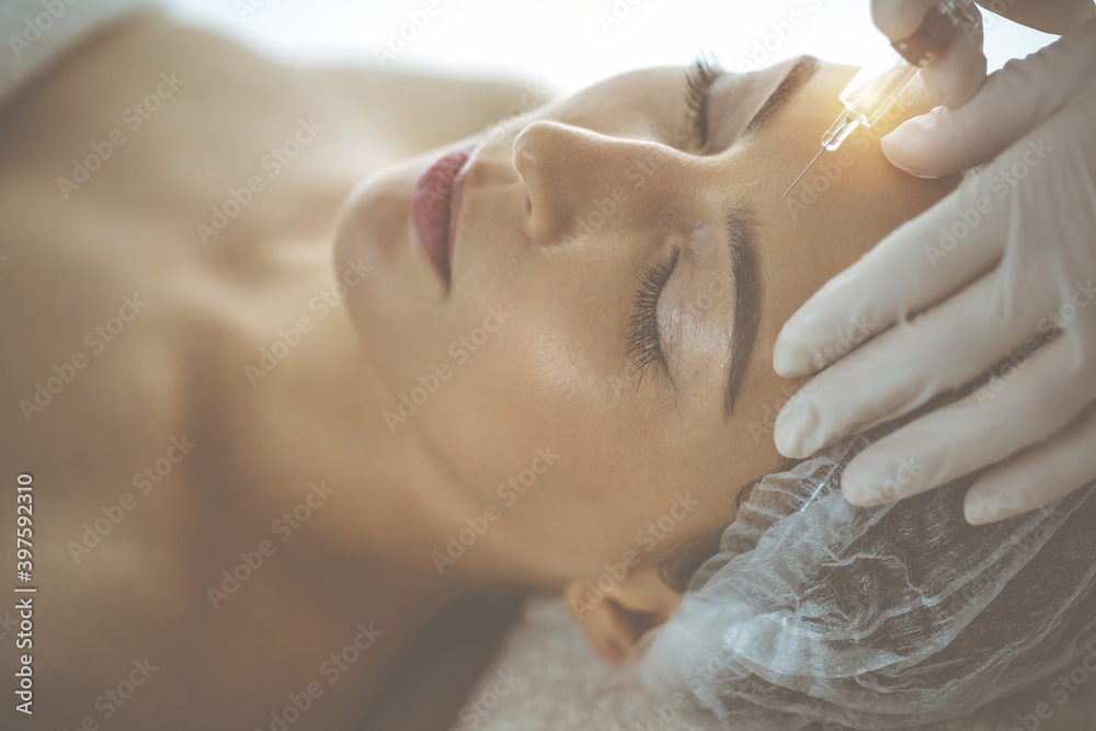 Beautician doing beauty procedure with syringe to face of young brunette woman in sunny clinic. Cosmetic medicine and surgery, beauty injections Beautician doing beauty procedure with syringe to face of young brunette woman in sunny clinic. Cosmetic medicine and surgery, beauty injections