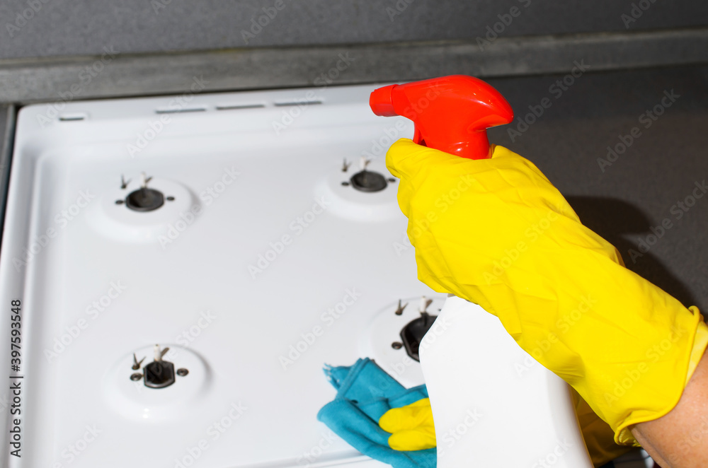 Female hand in rubber glove wiping the surface of gas stove, close-up. Woman washing kitchen apartment with cleaning spray