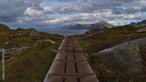 Wooden footbridge on hiking trail crossing muddy areas between grass and rocks on Moskenesøya island, Lofoten, Norway with rough mountains on the coast in background. Focus on footbridge.