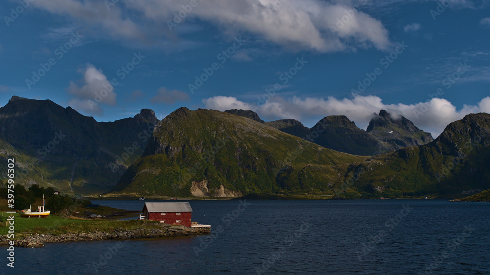Red painted small shed on the shore of fjord Selfjorden on Moskenesøya island, Lofoten, Norway with majestic mountains of Flakstadøya in background including rugged peak of Volandstinden.