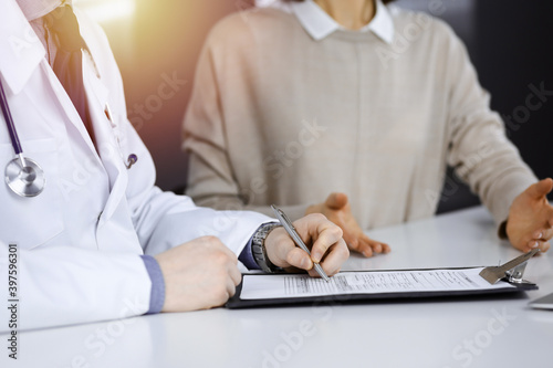 Unknown male doctor and patient woman discussing something while sittingin a darkened clinic and using clipboard, glare of light on the background photo