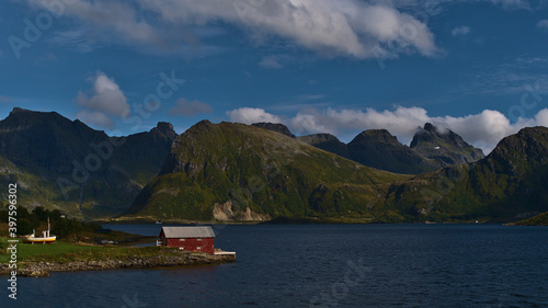 Red painted small shed on the shore of fjord Selfjorden on Moskenesøya island, Lofoten, Norway with majestic mountains of Flakstadøya in background including rugged peak of Volandstinden.