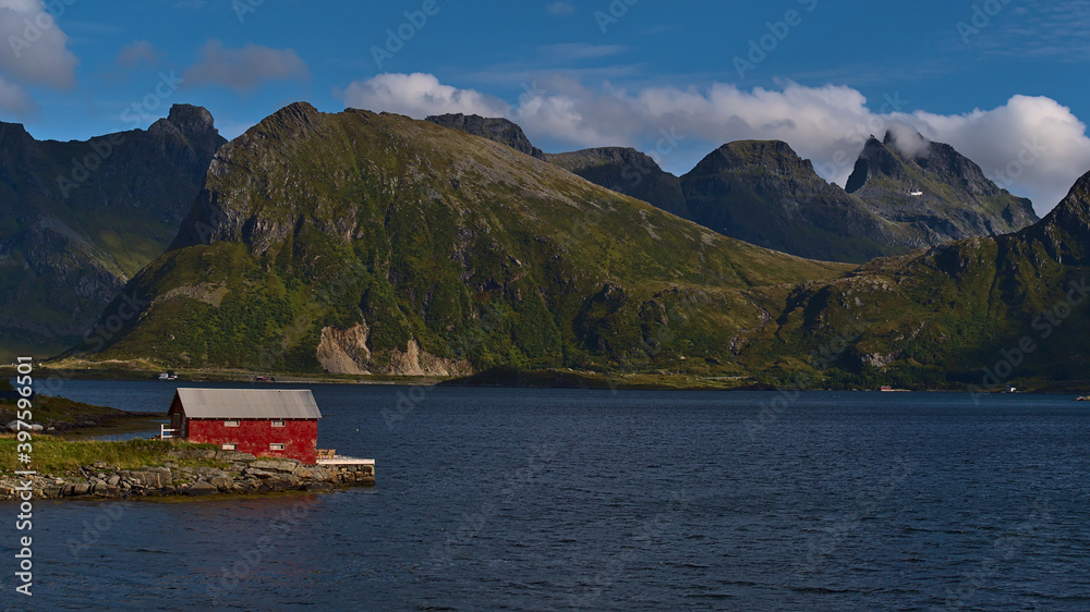 Idyllic view of small shed with red painted facade located on the shore of peaceful fjord Selfjorden with the stunning rugged mountains of Flakstadøya island in background on sunny day in late summer.