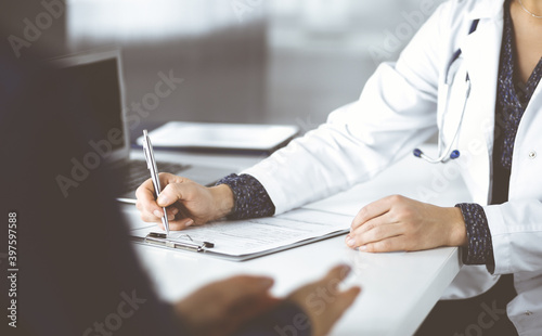 Unknown woman-doctor is writing some medical recommendations to her patient, while they are sitting together at the desk in the cabinet in a clinic. Physician is using a clipboard, close-up. Perfect