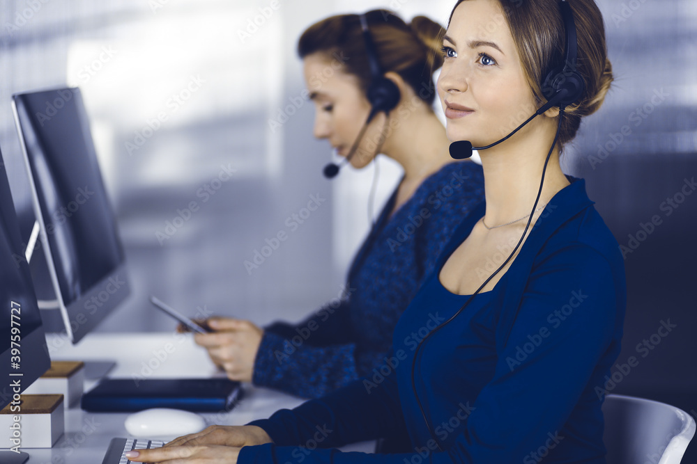 Two busineswomen have conversations with the clients by headsets, while sitting at the desk in a modern office. Diverse people group in a call center. Telemarketing and customer service Two busineswomen have conversations with the clients by headsets, while sitting at the desk in a modern office. Diverse people group in a call center. Telemarketing and customer service