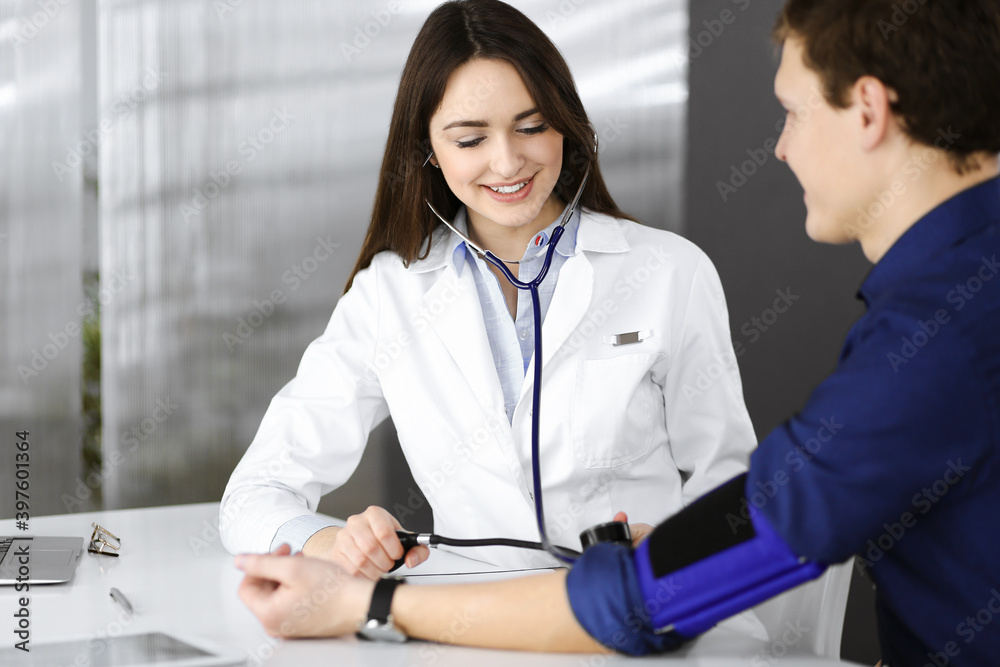 Young woman-doctor is checking her patient's blood pressure, while they are sitting together at the desk in a cabinet. Physician at work in a hospital. Medicine and healthcare concepts Young woman-doctor is checking her patient's blood pressure, while they are sitting together at the desk in a cabinet. Physician at work in a hospital. Medicine and healthcare concepts