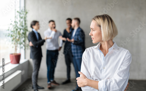 Male Coworkers Whispering Behind Back Of Unhappy Businesswoman In Office photo