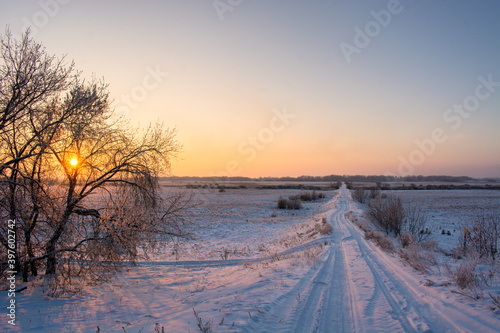 Winter rural, country road on hill in field covered by snow at sunset with the sun shining through the trees with sky gradient