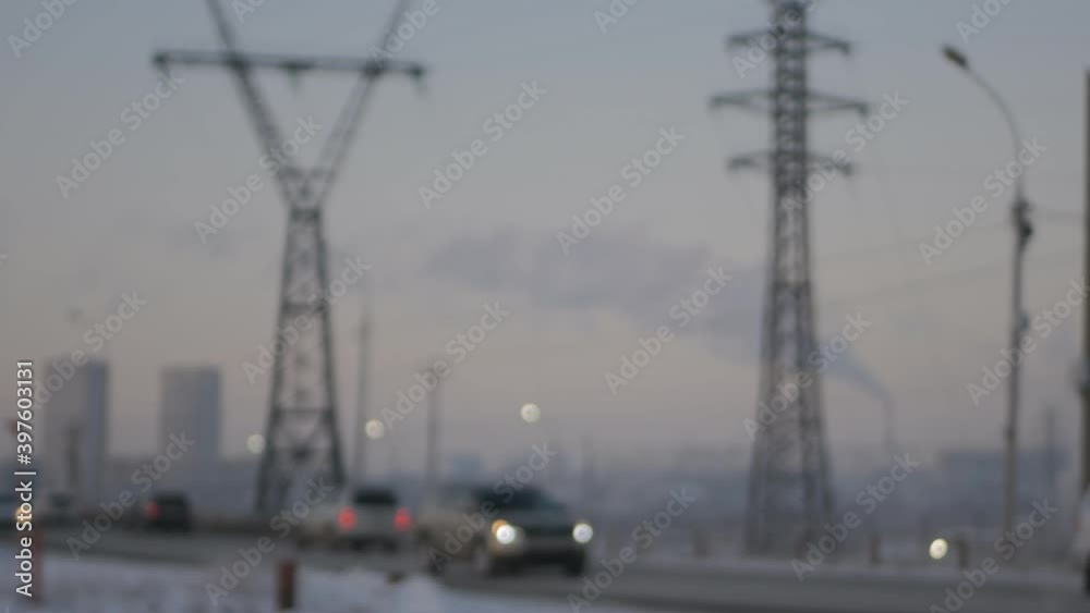 Blurred. Cars are driving along the road in the city. Supports visible High voltage electricity lines. Smoke goes from the chimney to the sky. Industrial view of the city