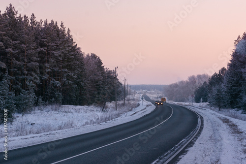 Turn of curved asphalt road with car traffic  with shining lights in distance running through winter pine forest covered with frost at sunset with pink sky gradient in blue hour © Денис Кузнецов
