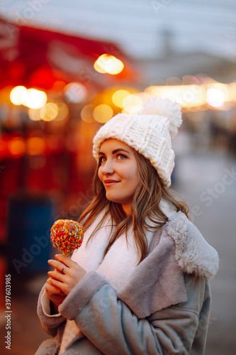 Happy woman with caramel apple on Festive Christmas fair. Beautiful woman enjoys winter holidays on Christmas market. New year.Lights around.