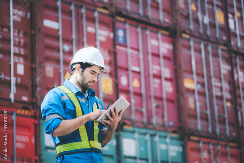 Man wears hardhat and reflection shirt and checking tablet with blurry metal containers in background. Concept of inventory and logistic management.