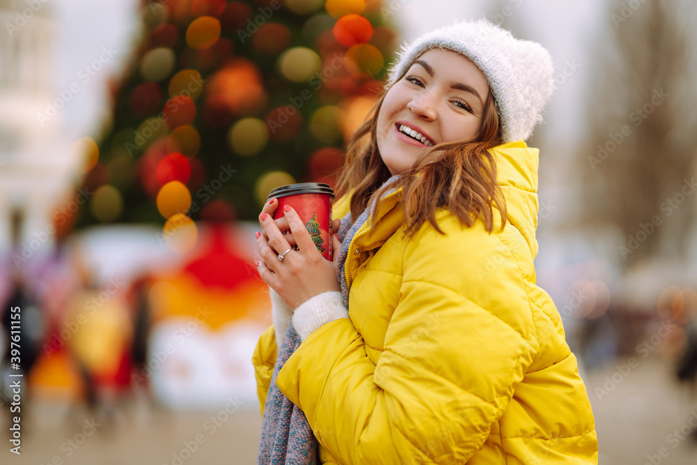 Happy woman drinking hot coffee while walking near New year tree on Festive fair. Cold weather. Winter fashion, holidays, rest, travel concept. Happy woman drinking hot coffee while walking near New year tree on Festive fair. Cold weather. Winter fashion, holidays, rest, travel concept.