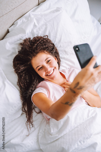 Young smiling afro hair woman lie in the bed and use smartphone making selfie. Social media addiction, online dating.