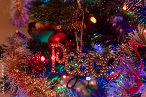A close up of a Christmas Tree ornament of the word "peace", hanging on a tree among the tinsel, multicoloured LED lights, and other ornaments.