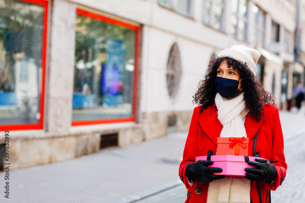 Portrait of a woman in red shopping outside with face mask holding many christmas gifts and presents looking at shop windows